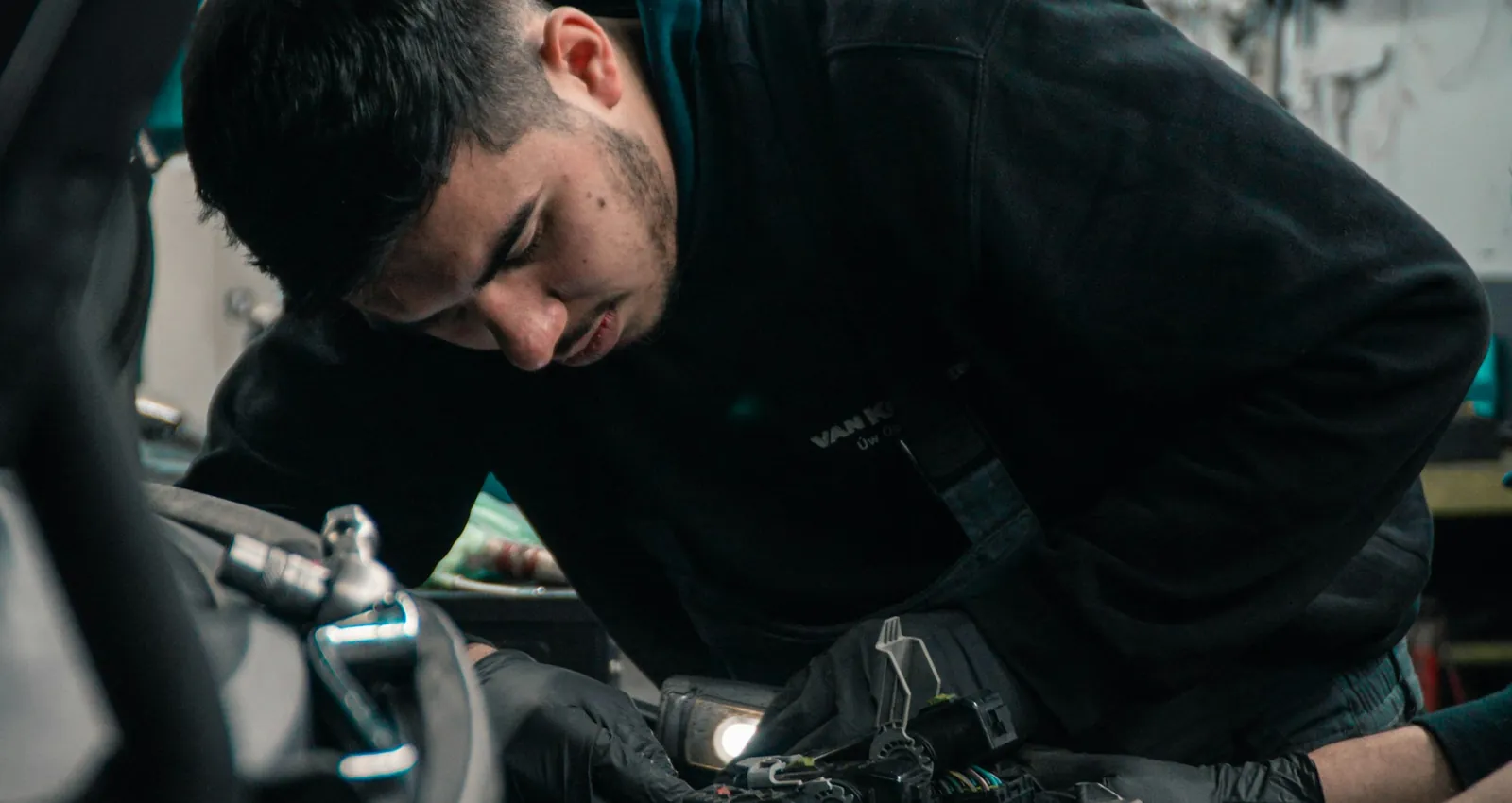 Technician in black gloves diagnosing engine wiring under the hood of a car in a European repair shop